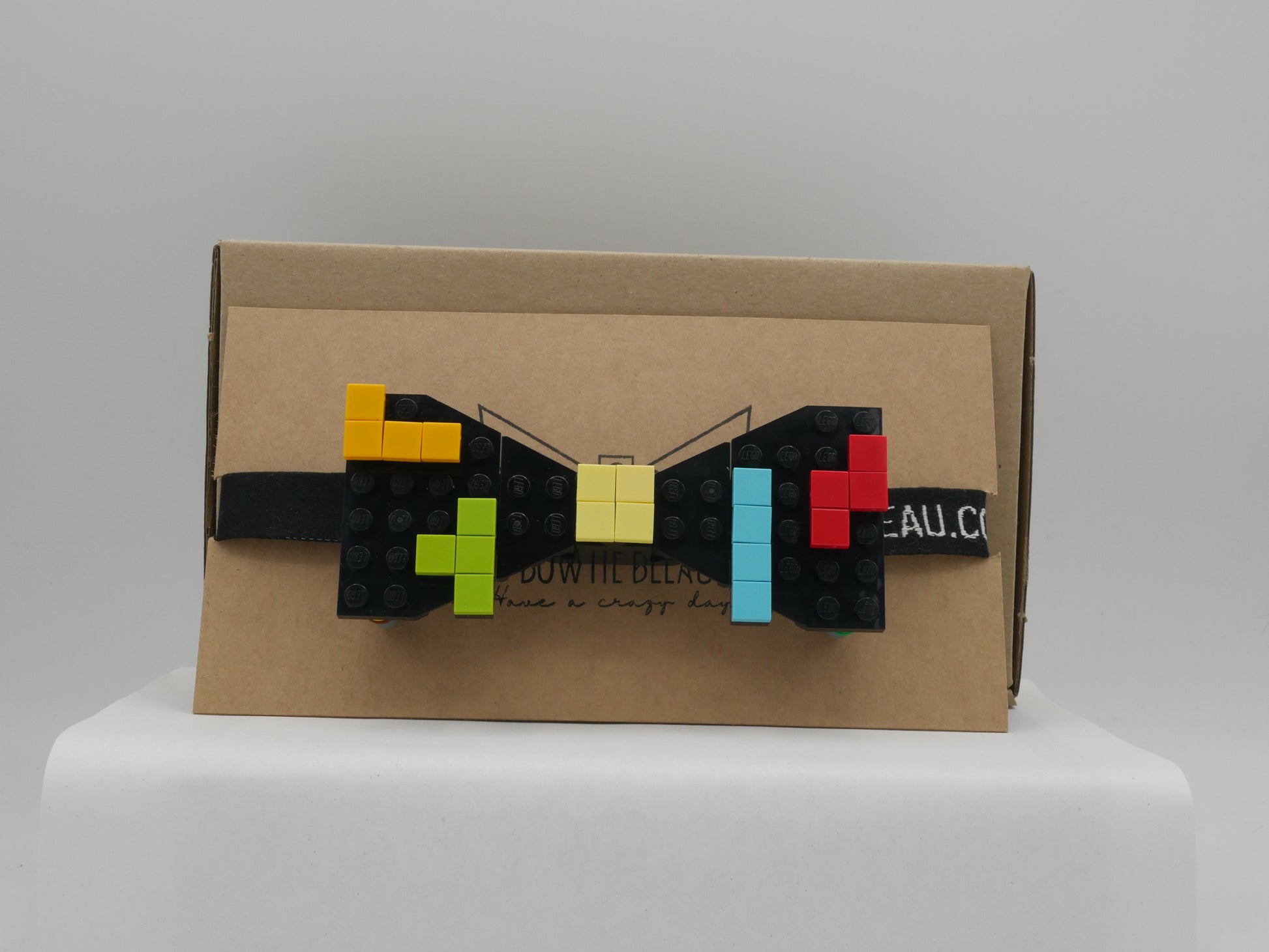 A black LEGO® bow tie with a retro arcade design featuring yellow, orange, green, red, and blue blocks, displayed on a cardboard box.