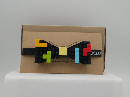 A black LEGO® bow tie with a retro arcade design featuring yellow, orange, green, red, and blue blocks, displayed on a cardboard box.