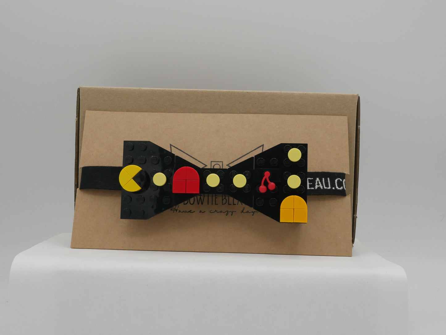 A black LEGO® Retro Arcade Maze Bow Tie with a strap, featuring yellow, orange, red, and cherry LEGO® parts on a cardboard box.