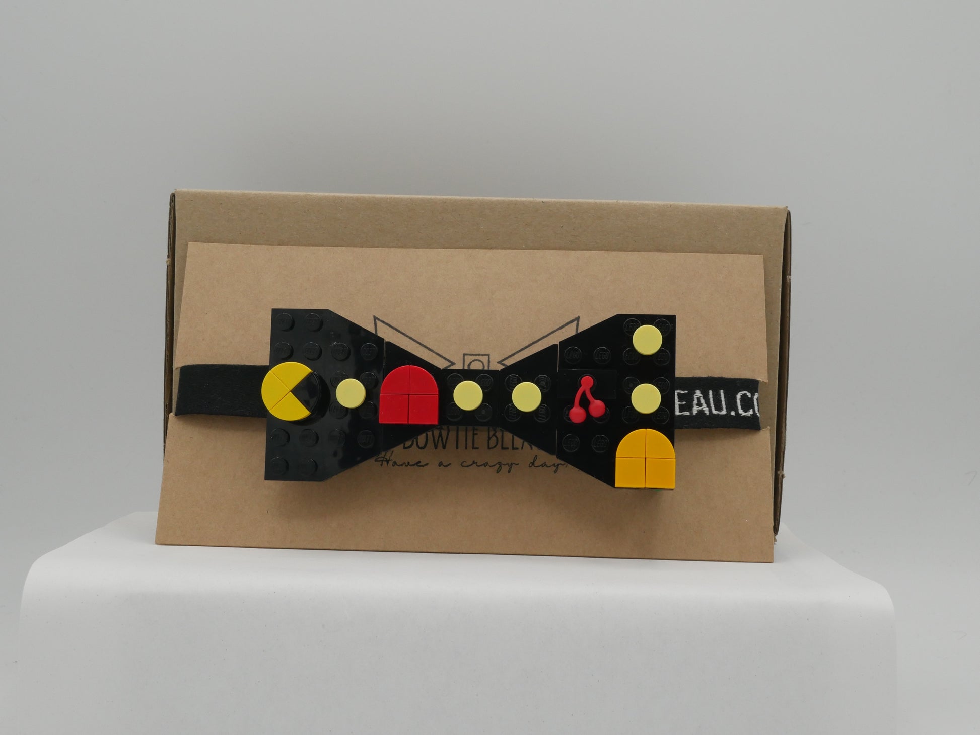 A black LEGO® Retro Arcade Maze Bow Tie with a strap, featuring yellow, orange, red, and cherry LEGO® parts on a cardboard box.